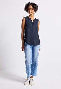 Granatowa bezrękawnik z dwoma kieszeniami na piersi, dekoltem w serek i bocznymi rozporkami, w połączeniu z jasnoniebieskimi prostymi dżinsami oraz białymi trampkami.