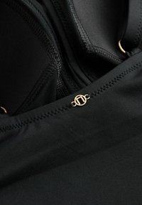 Kratki prikaz crnog kostima za kupanje s mrežastim detaljima i malim zlatnim kružnim logom na prednjem središnjem šavu.