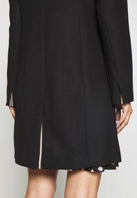 Black tailored coat with a textured finish, side slits, and a subtle light-colored lining visible at the cuffs and hem.