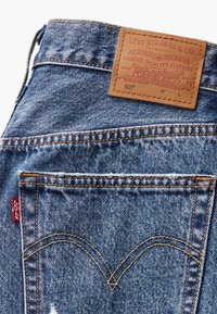 Plave traperice s kožnim oznakom brenda, narančastim šavovima i crvenom Levi's oznakom. Imaju klasičan dizajn stražnjeg džepa u obliku slova V.