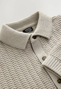 Polo en tricot beige avec un col, deux boutons foncés et un motif texturé. Le matériau semble doux et chaud.
