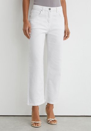 Femme portant un jean blanc taille haute à jambe droite avec bouton et poches sur le devant, associé à des sandales à talons hauts beiges à brides.