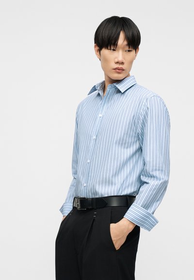 Νεαρός άνδρας με μπλε και λευκό ριγέ πουκάμισο με κουμπιά και μαύρο παντελόνι με μαύρη ζώνη, με τα χέρια στις τσέπες, κοιτάζει στο πλάι μπροστά σε απλό φόντο.