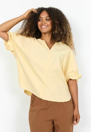 Donna sorridente con capelli ricci, indossa una camicetta larga di colore giallo pallido e pantaloni marroni, posa con una mano nei capelli su sfondo bianco.