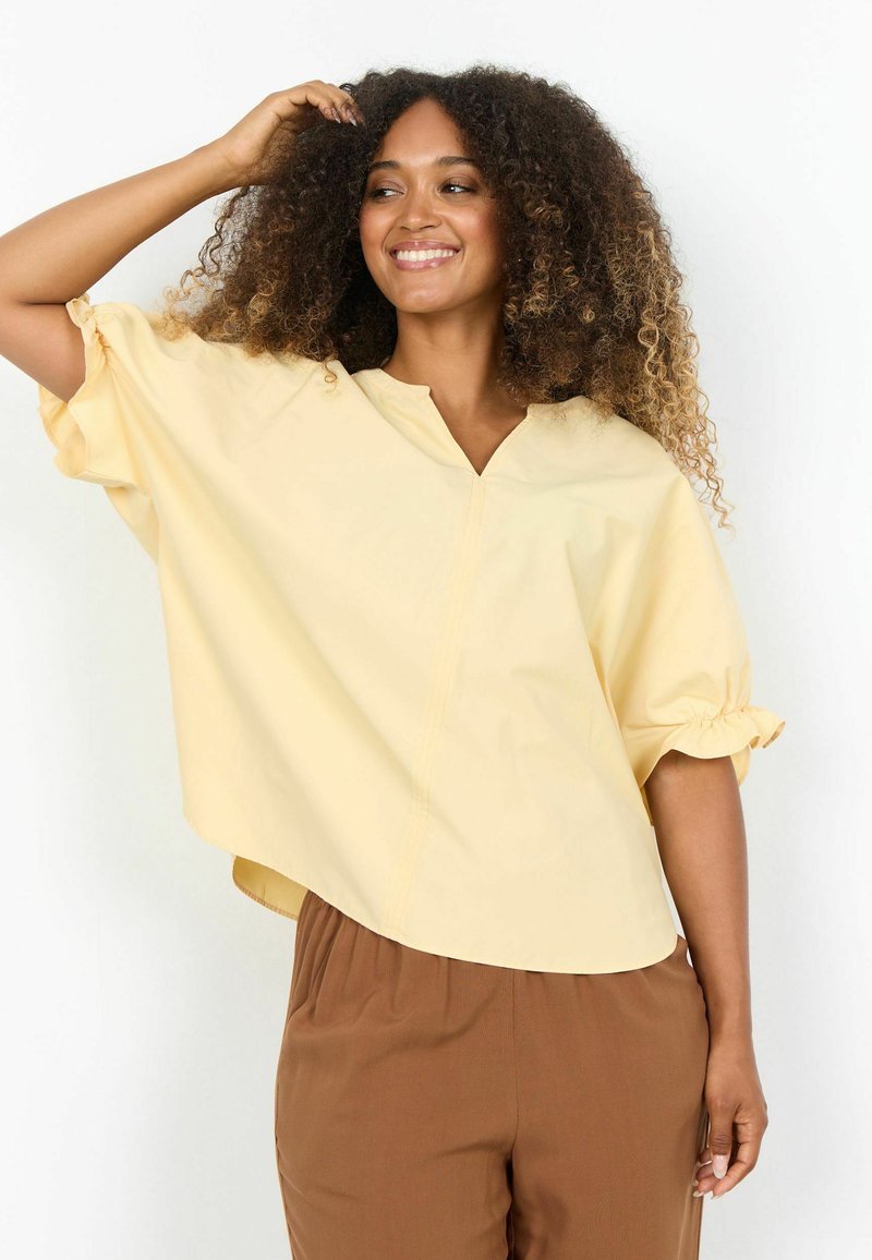 Femme souriante aux cheveux bouclés portant un chemisier ample jaune pâle et un pantalon marron, posant avec une main dans les cheveux sur un fond blanc.