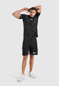 Camiseta atlética negra con logo, combinada con pantalones cortos negros que presentan un acento blanco. El modelo lleva zapatillas blancas, de pie en una pose neutral.