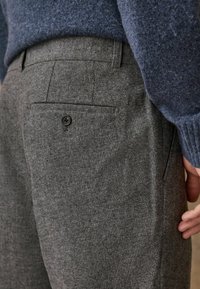 Gray textured trousers with a subtle herringbone pattern, featuring a back pocket with a black button, worn with a blue knitted sweater.