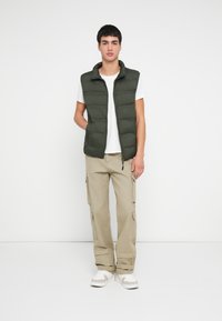 Mladý muž stojaci s jednou rukou v kapsách olivovo zelenej nadmernej vetrovky, biele tričko, béžové cargo nohavice a biele tenisky na jednotvárnom pozadí.