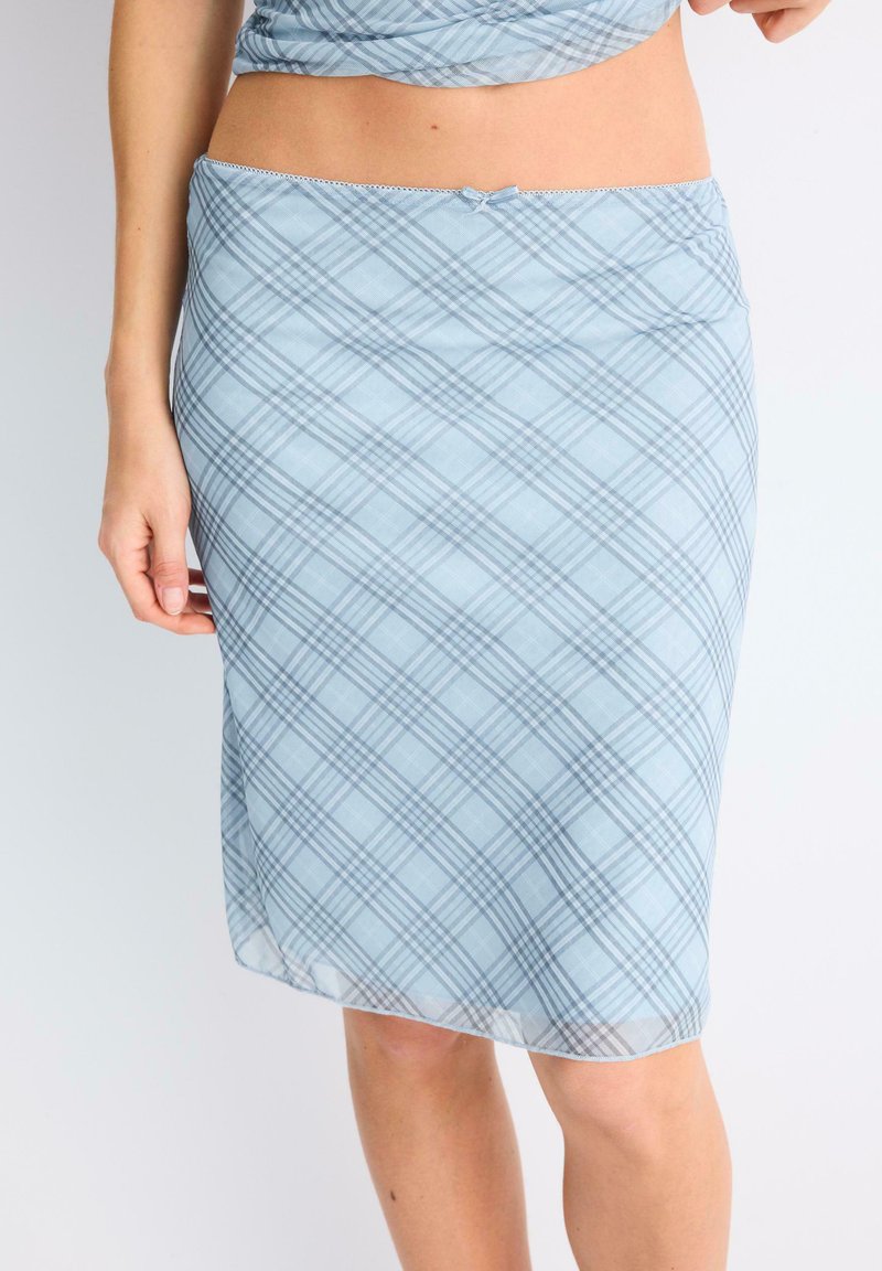 Femme portant une jupe à carreaux bleu clair jusqu'aux genoux et un haut assorti, une main reposant le long du corps sur un fond uni.