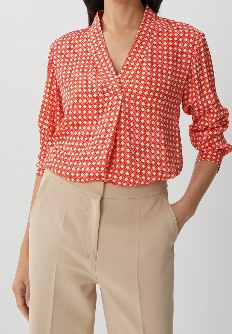 Femme portant un chemisier rouge et blanc à motifs avec des manches longues et un pantalon beige taille haute, une main dans la poche.