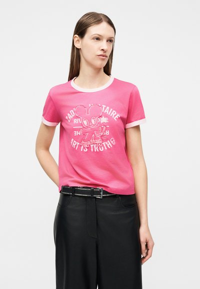 Γυναίκα που φοράει ροζ κοντομάνικη μπλούζα με λευκό περίγραμμα και γραφικό με καρτούν ποντίκι που κάνει σκέιτμπορντ, σε συνδυασμό με μαύρο παντελόνι και ζώνη.