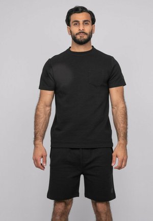 Homme barbu portant une chemise noire à manches courtes texturée et un short noir assorti, debout avec une expression neutre sur un fond uni.
