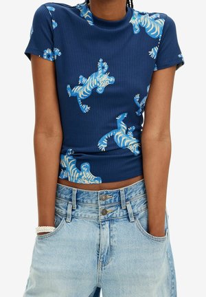 Kobieta ubrana w dopasowaną prążkowaną granatową bluzkę z krótkim rękawem z niebiesko-białymi nadrukami zebry oraz jasnoniebieskie dżinsy z podwójnym zapięciem na guziki.