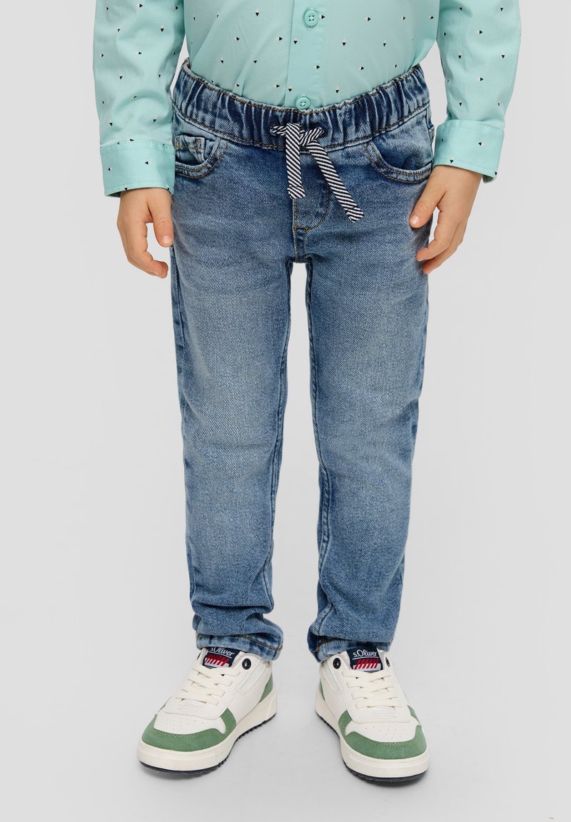 Enfant portant une chemise bleu clair à motif, un jean bleu à taille élastique avec cordon rayé, et des baskets blanches avec des touches de vert.
