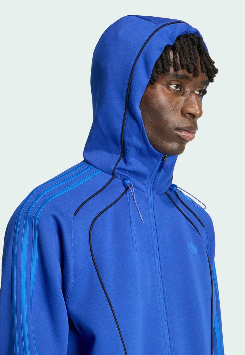 Homme faisant face à droite portant une veste à capuche bleu vif avec des bordures noires et trois bandes bleu clair sur les manches.