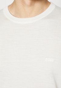 Suéter de punto color crema con un escote redondo y una textura suave. Presenta un pequeño logo blanco de "BOSS" en el pecho.