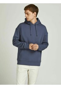 Dark blue hoodie made of soft fabric, featuring a front pocket, adjustable drawstring hood, and ribbed cuffs. Worn with light-colored pants.
