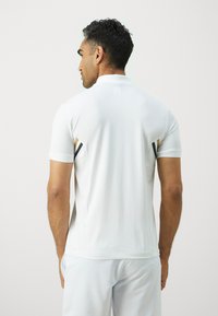 White polo shirt with short, textured sleeves, contrasting black and gold accents on the shoulders. Soft, smooth fabric and fitted design.