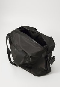 Svart stoff skulderveske med glidelåser, justerbar stropp og en glatt tekstur. Har et fremtredende logo langs siden.