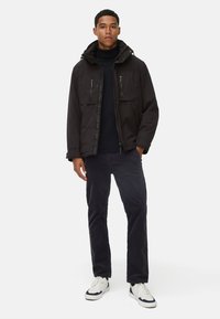 Schwarze gepolsterte Jacke mit Kapuze, Frontreißverschluss, zwei Brusttaschen und wasserabweisender Oberfläche, getragen über einem schwarzen Rollkragenpullover und dunklen Hosen.