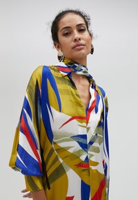 Abito colorato, lungo fino al ginocchio, realizzato in tessuto morbido; presenta audaci motivi astratti in giallo, blu, verde, rosso e bianco; con colletto e sciarpa.