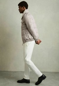 Jachetă puffer de culoare gri deschis, cu textura matlasată, cu manșete cu nervuri, pantaloni albi slim și cizme negre până la gleznă. Modelul stă în profil.