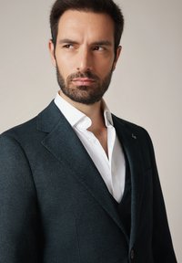 Mężczyzna z ciemnymi włosami i brodą, ubrany w ciemną, teksturowaną marynarkę i białą koszulę, spoglądający na bok na neutralnym tle.