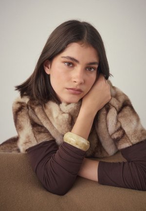 Brown faux fur wrap with a layered texture, worn over a fitted brown long-sleeve top. An opaque light yellow bangle adorns the wrist.