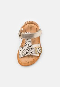 Lapsen beige nahkasaapas, jossa on mustia pilkkuja. Varustettu kullanvärisellä soljella ja vaaleanruskealla, lehtikuviollisella pohjallisella. Avoin varvasmuoto.