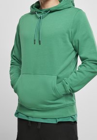 Grüner Hoodie aus weichem Material. Verfügt über eine Kapuze mit Kordelzug, eine Fronttasche und gerippte Bündchen mit lockerem Schnitt.
