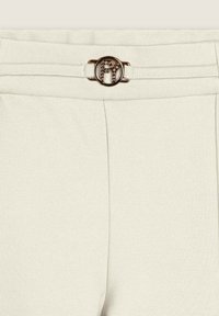 Pantalones beige con una textura suave, que cuentan con una hebilla circular plateada con un logotipo y piedras decorativas en la pretina.