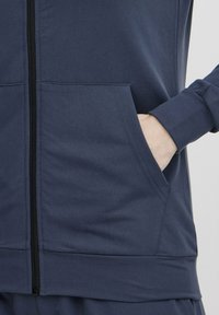 Navy zipsvārku džemperis ar augstu apkakli, priekšējām kabatām un ribotām aprocēm. Izgatavots no gludas, elastīgas auduma ar tīru apdari.