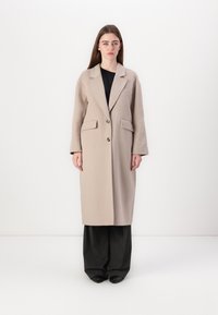 Beige villasekoite takki, jossa on kauluskanta, kaksirivinen nappisulku ja kaksi etutaskua. Muotoilu on suora ja siinä on pitkät hihat.
