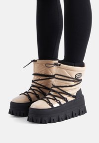 Beżowe, puchate botki do kostki z grubą, czarną podeszwą. Posiadają czarne sznurówki i akcenty, błyszczące wykończenie oraz zaokrąglony kształt noska.