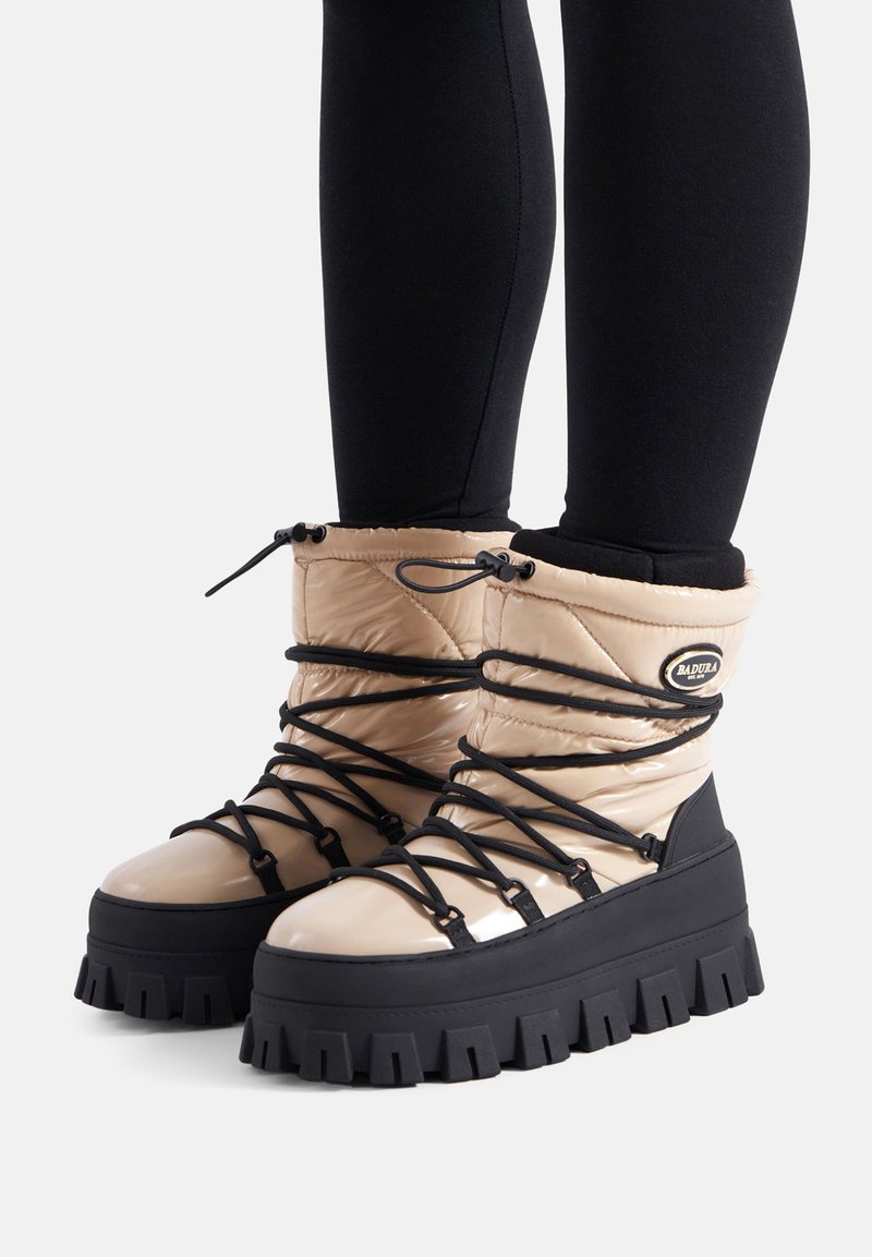Beżowe, puchate botki do kostki z grubą, czarną podeszwą. Posiadają czarne sznurówki i akcenty, błyszczące wykończenie oraz zaokrąglony kształt noska.