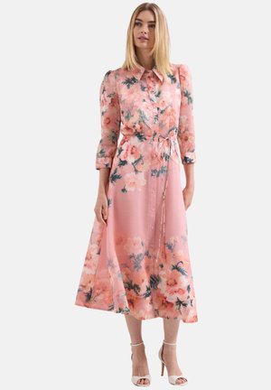 Femme portant une robe midi rose à fleurs à manches longues avec des boutons sur le devant, taille nouée, et des sandales blanches à talons hauts, debout.