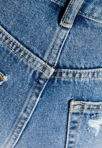 Denimtyg med en blå nyans, med gul överstygnssöm. Närbild av fickor och sömmar som visar textur och lätt slitage.