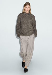 Overmaatse donkerbruine gebreide trui met een hoge hals, gecombineerd met lichtgrijze geplooide broeken en zwarte instappers.