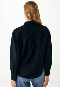 Czarna koszula z długim rękawem o luźnym kroju, z detalicznym przeszyciem, spiczastym kołnierzykiem i zapięciem na guziki, zestawiona z niebieskimi dżinsami.