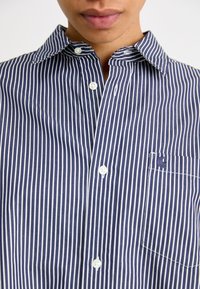 Chemise rayée bleu marine et blanche avec un col boutonné, une poche avant avec un logo brodé et des boutons blancs.