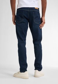 Jeans de mezclilla azul oscuro con un ajuste slim, bolsillos traseros con costuras sutiles, y combinados con zapatillas de color claro.