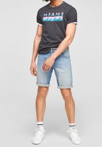 Harma t-paita, jossa on "MIAMI VICE" -graafikka, vaaleansiniset kääntödenim-shortsit, valkoiset lenkkarit ja raidalliset sukat. Rentoa, löysää istuvuutta.