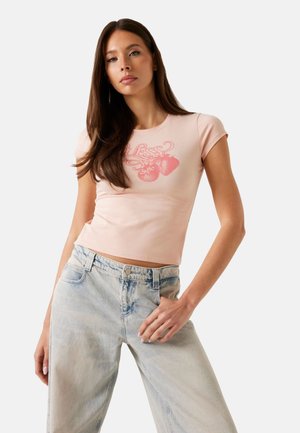 T-shirt fitted rosa chiaro con maniche corte e scollo rotondo, con una grafica di fragola rossa. Indossato con jeans di denim blu chiaro.