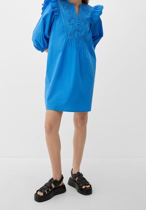 Femme portant une robe bleu vif jusqu'aux genoux avec des manches bouffantes et des broderies, associée à des sandales noires à plateforme.