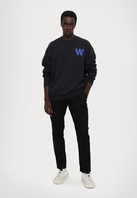 Svart genser med blå logo på venstre bryst, kombinert med svarte bukser og hvite sneakers. Glatt stoff, avslappet passform.