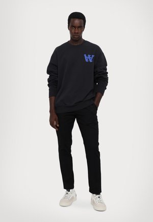 Zwarte sweatshirt met blauw logo op de linkerborst, gecombineerd met zwarte broek en witte sneakers. Gladde stof, ontspannen pasvorm.