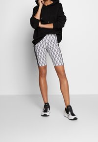 Svart hoodie med dragsnoddar, tillsammans med vita mönstrade cykelbyxor med geometriska mönster och svarta sidostripes. Svarta sneakers.