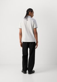 Weißes Polohemd mit marineblauen Kragen- und Ärmelakzenten, kombiniert mit schwarzen Hosen und schwarzen Schuhen, von hinten betrachtet.