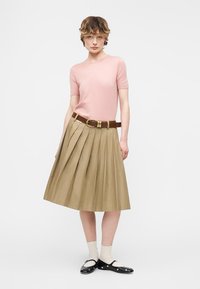 Νεαρό άτομο που φοράει ροζ κοντομάνικη μπλούζα, μπεζ πλισέ φούστα με καφέ ζώνη, λευκές κάλτσες και μαύρα φλατ παπούτσια με στολίδια, όρθιο.