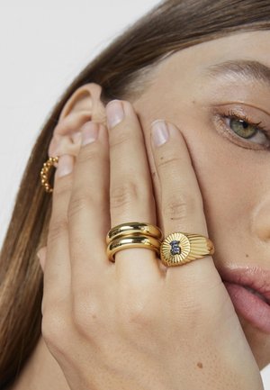 Anelli d'oro su una mano: due bande lisce e una con un design a raggiera testurizzato che presenta una gemma centrale. La pelle è di tonalità chiara e non lucida.